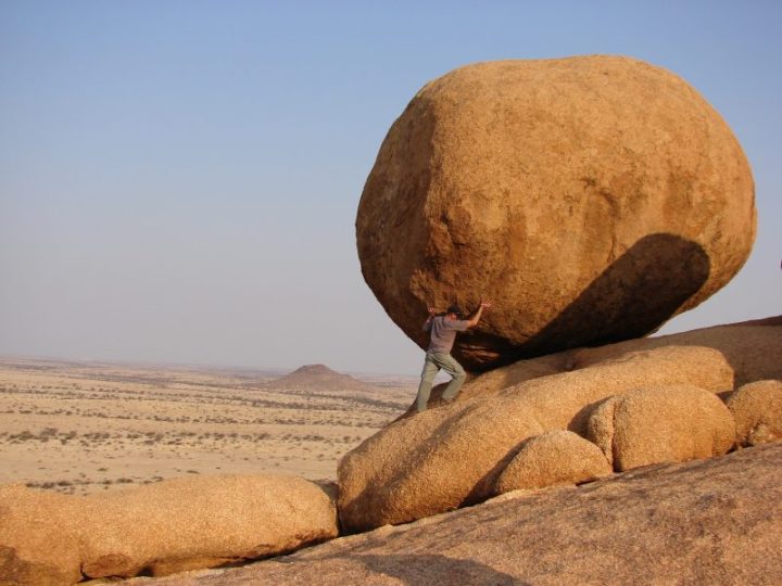 Man pretending to 'hold up' a large boulder on a desert-like plain.